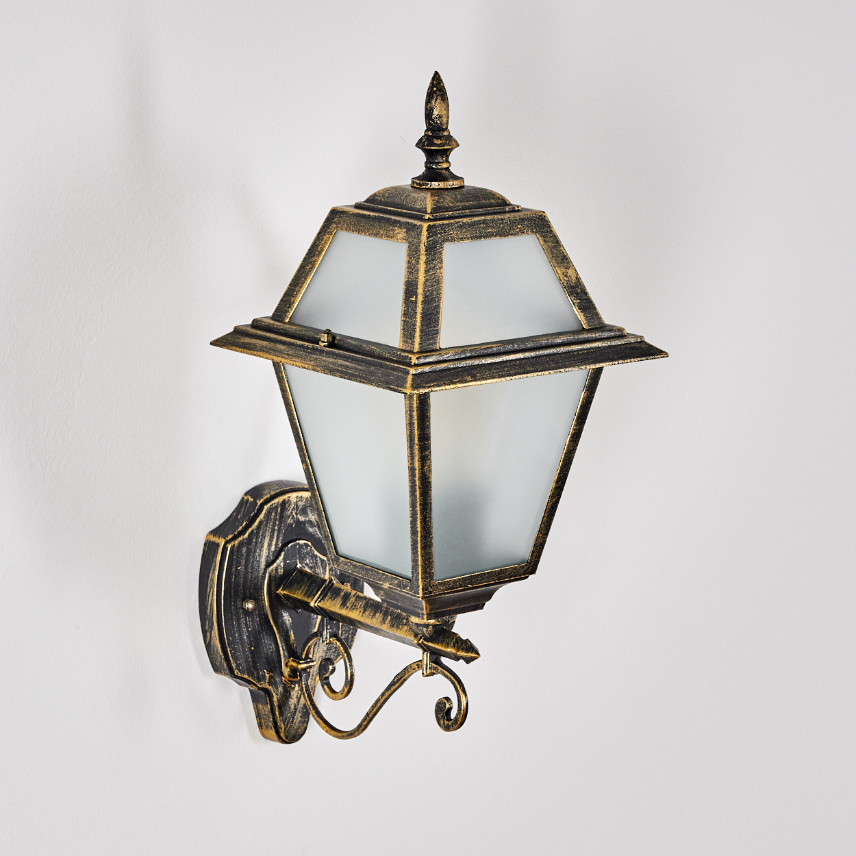Eine verzierte Wandleuchte mit einem Schirm aus Milchglas. Sie hat eine schwarz-goldene Oberfläche an einer weißen Wand.