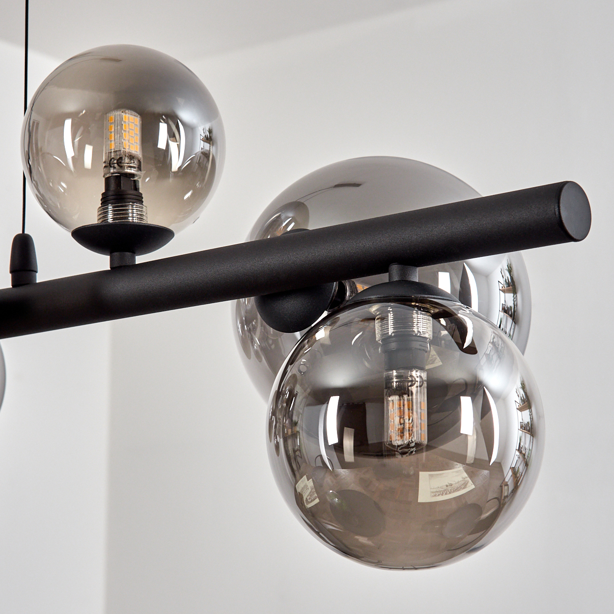 Eine moderne schwarze Pendelleuchte mit vier rauchfarbenen Glaskugeln, jede mit einer sichtbaren Glühbirne, vor einer weißen Wand.