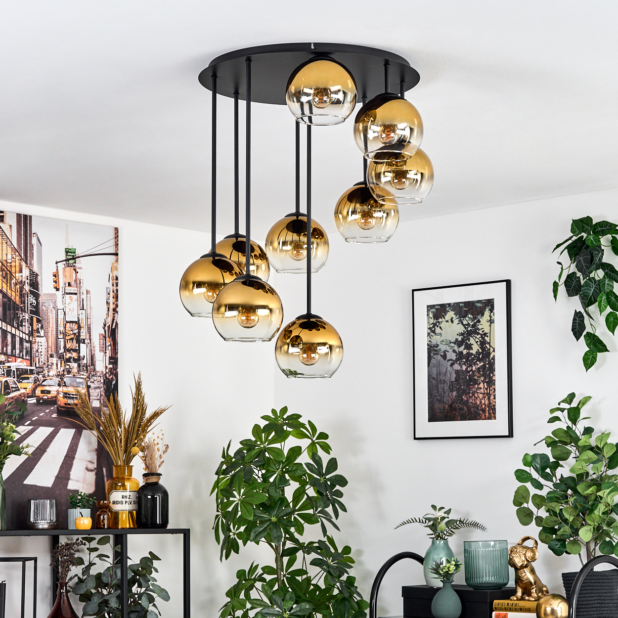 Eine moderne Deckenleuchte mit goldenen Glaskugeln. Grüne Pflanzen und Kunstwerke schmücken die weiße Wand. Schwarze Möbel und Dekoration sind sichtbar.