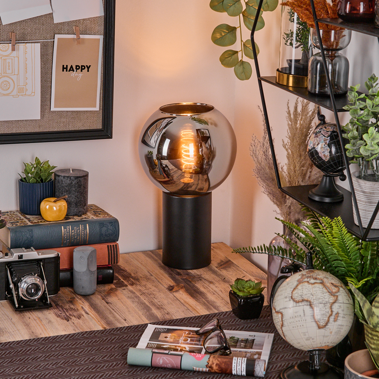 Ein Schreibtisch mit Lampe, Büchern, Kamera, Globus und Pflanzen. Ein 'Happy Day'-Notiz an der Wand.