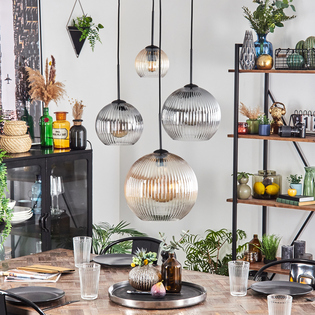 Esszimmer mit Hängelampen, Tisch mit Gläsern und Tellern sowie Dekorationsartikeln auf Regalen und Schränken.