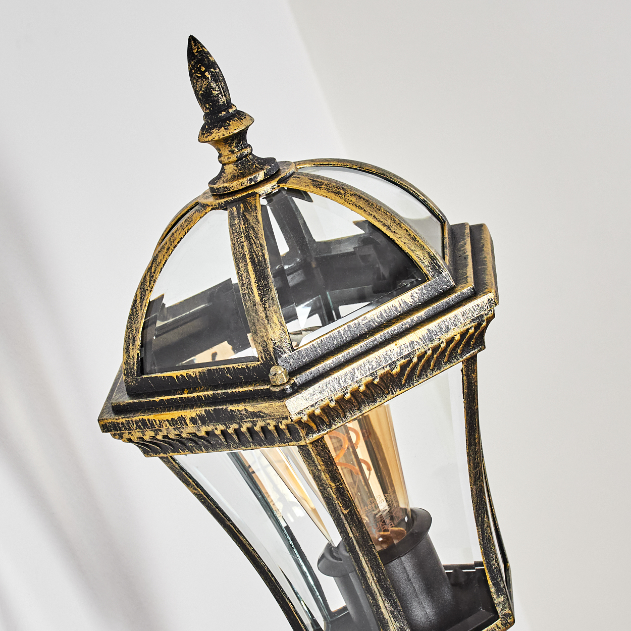 Nahaufnahme einer Vintage-Außenlaterne mit Gold- und Schwarzakzenten. Die Glasscheiben umgeben eine Glühbirne, vor einer weißen Wand.