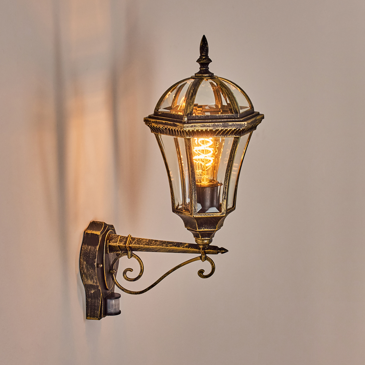 Eine Außenwandleuchte mit dekorativem Design. Sie hat ein Glasgehäuse und eine warme Glühbirne. Die Wand ist hellbeige.