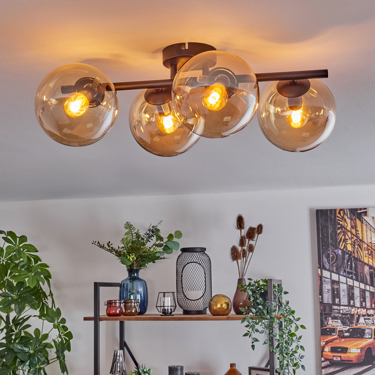 Deckenlampe mit vier bernsteinfarbenen Glaskugeln. Darunter ein Regal mit Deko: Pflanzen, Vasen und ein Taxibild.