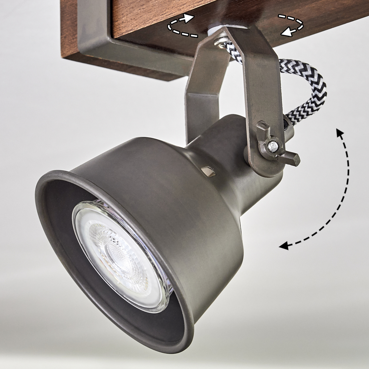 Ein dunkelgrauer Scheinwerfer ist auf einer Holzbasis montiert. Pfeile zeigen verstellbare Winkel an. Ein schwarz-weißes Kabel ist sichtbar.