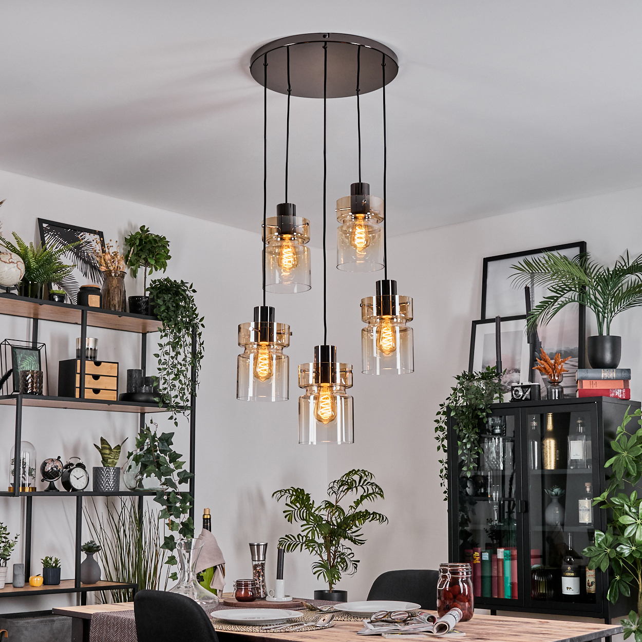 Esszimmer mit Kronleuchter. Tisch mit Tellern, Gläsern und Stühlen. Regale und ein Schrank mit Pflanzen.