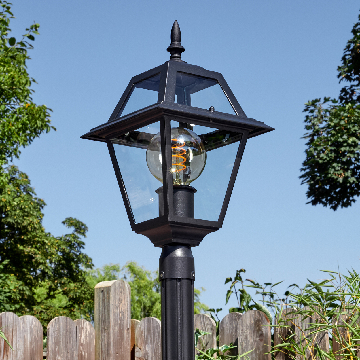 Schwarze Außenlampe mit Glasscheiben und einer dekorativen Glühbirne. Vor blauem Himmel, Holzzaun und grünem Laub.