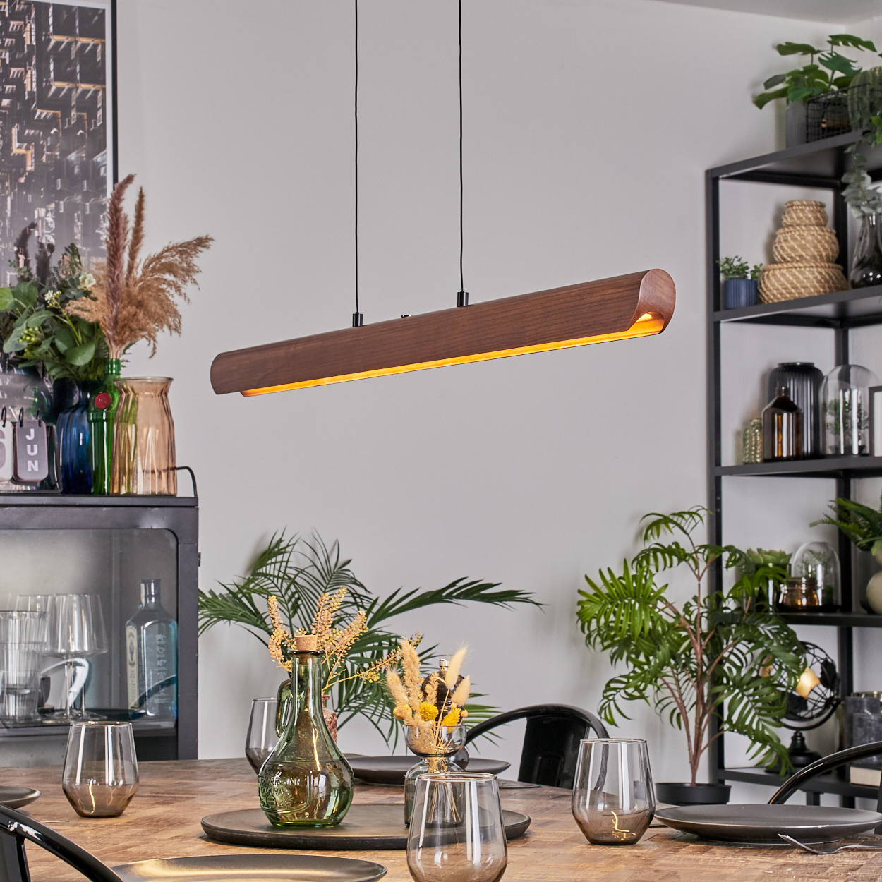 Esszimmer mit Holztisch, Gläsern, Pflanzen und einer langen Holzlampe. Regale mit Dekoration im Hintergrund.