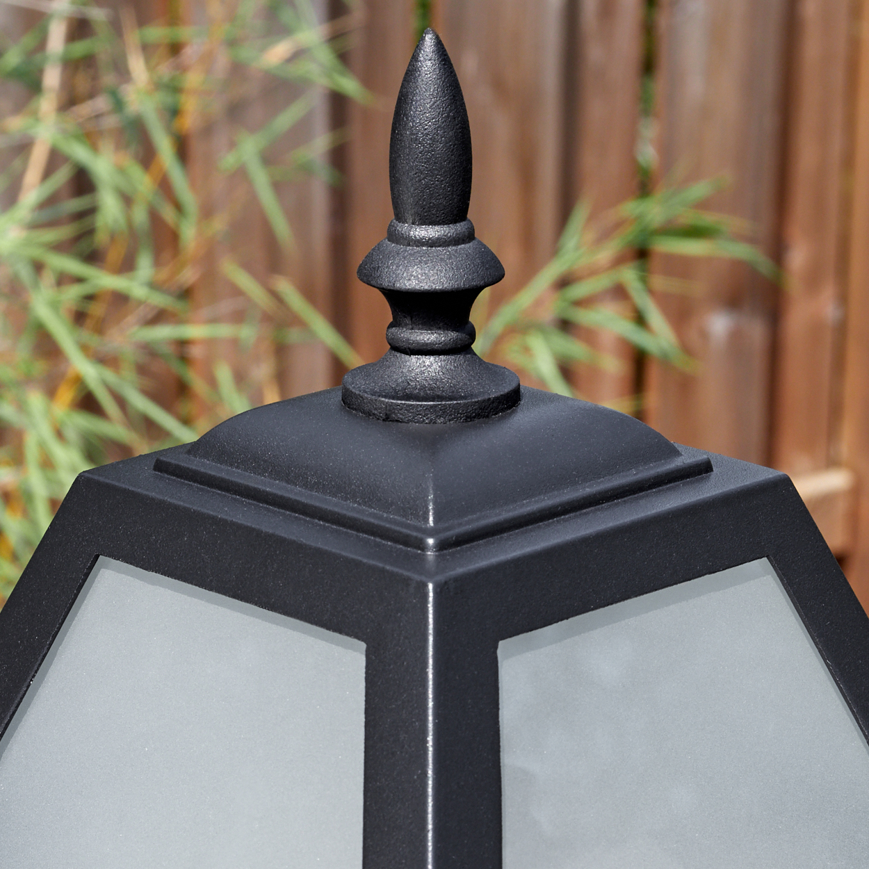 Nahaufnahme einer schwarzen Außenlampe mit einer matten Glasscheibe. Oben sitzt eine dekorative Spitze. Bambus und Holzzaun im Hintergrund.