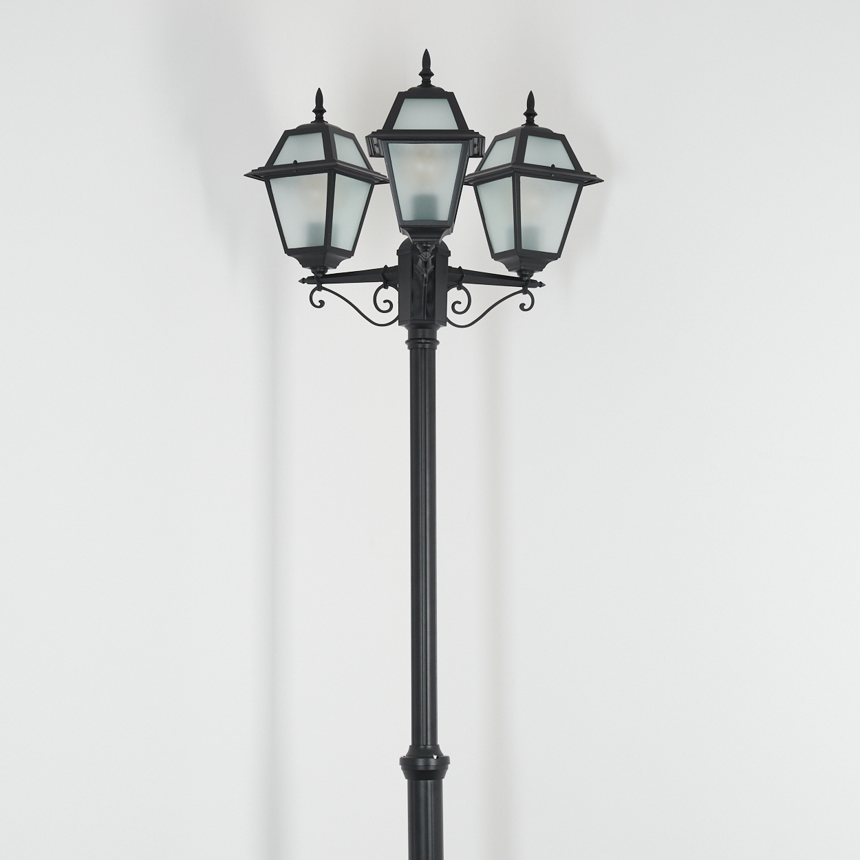 Eine schwarze Straßenlaterne mit drei Lampen steht vor einer weißen Wand. Die Lampen haben Milchglas und sind eingeschaltet.