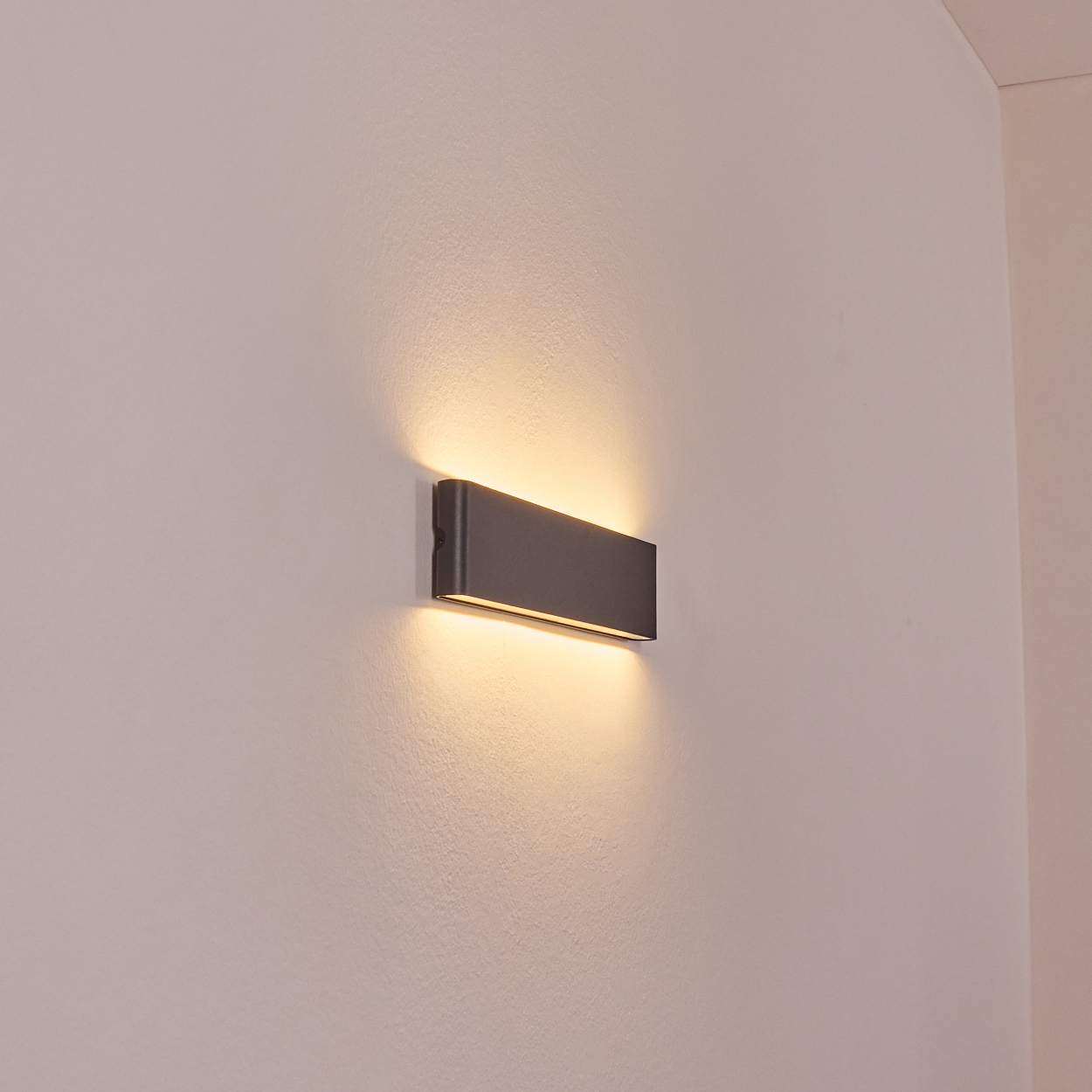Eine rechteckige, dunkelgraue Wandleuchte ist an einer weißen Wand befestigt. Die Lampe strahlt warmes, gelbes Licht aus.