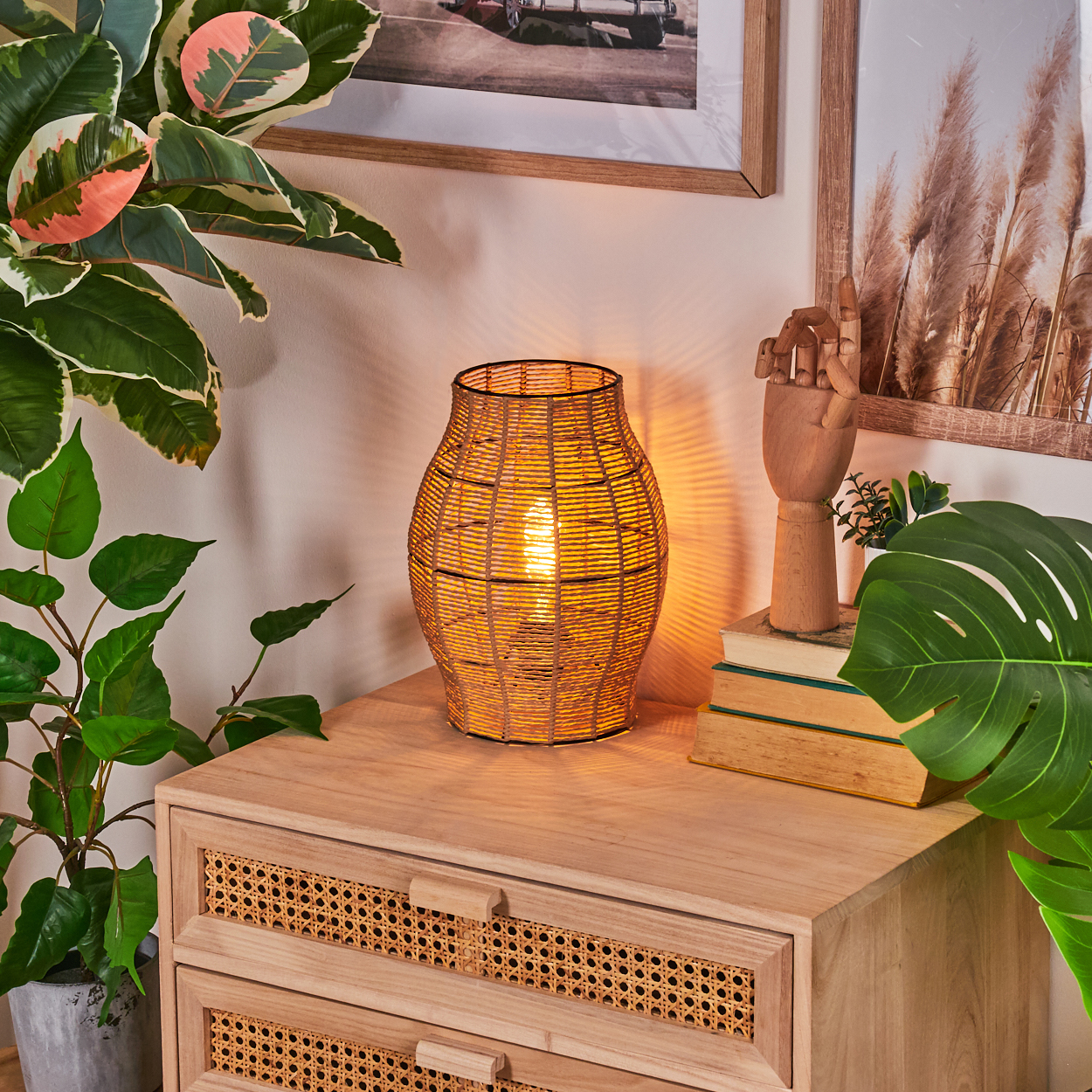 Eine beleuchtete gewebte Lampe steht auf einem Holzschrank, mit einer Holzhand, Büchern und Pflanzen. Bilder hängen an der Wand, und weitere Pflanzen flankieren den Schrank.