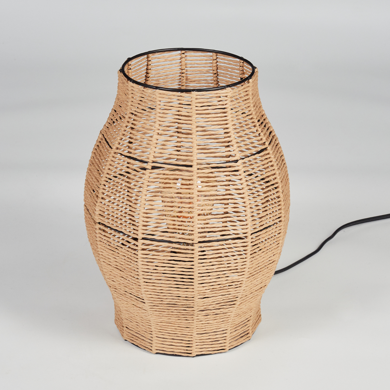 Eine gewebte, beige Lampe mit schwarzem Rand und schwarzen Stützstreifen. Ein schwarzes Kabel geht von der Lampe aus.