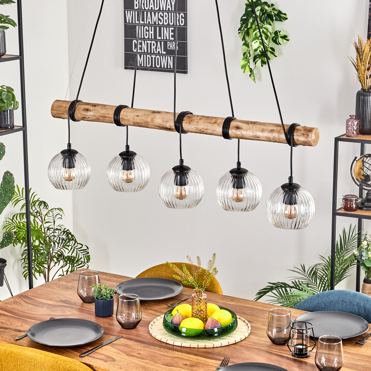 Ein Esstisch mit Tellern, Gläsern und einem Obst-Mittelstück. Ein Holzbalken mit Hängelampen beleuchtet die Szene.