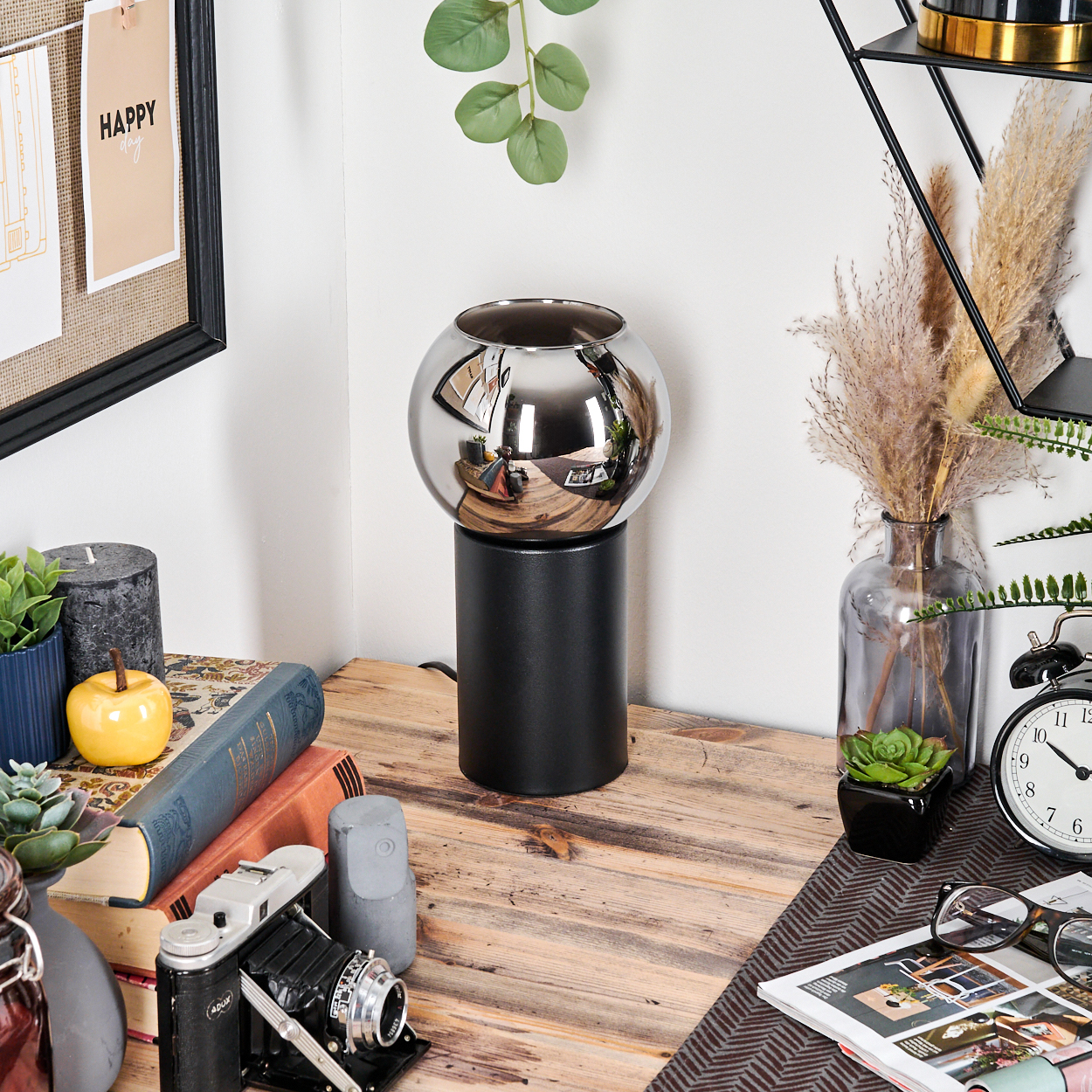 Eine Chromlampe steht auf einem Holztisch mit Büchern, einer Kamera und einer Uhr. Eine Tafel mit 'HAPPY day' ist an der Wand.