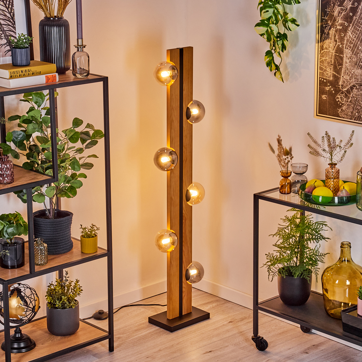 Eine moderne Stehlampe mit Glaskugeln steht neben Regalen mit Pflanzen und Dekoration. Im Hintergrund hängt eine Landkarte.