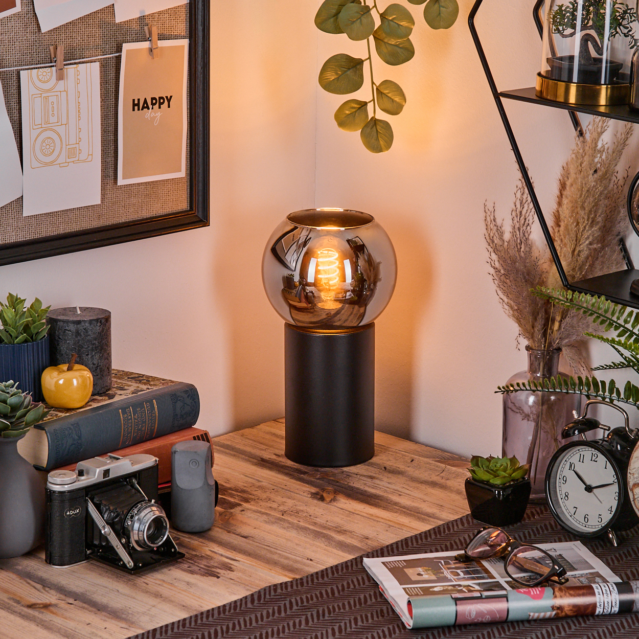 Eine Schreibtischecke mit Lampe, Büchern, Kamera und Dekoration. Ein 'Happy Day'-Zettel ist an einer Tafel befestigt.