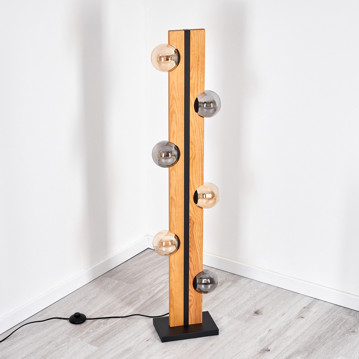 Eine moderne Stehlampe mit Holzrahmen und Glaskugeln. Die Lampe ist hoch und steht auf einem schwarzen Sockel. Der Hintergrund ist eine weiße Ecke.