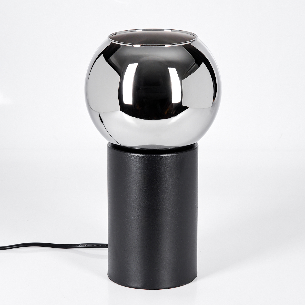 Eine moderne Lampe mit einer Chromkugel auf einem schwarzen zylindrischen Sockel. Ein schwarzes Kabel erstreckt sich von der Basis vor weißem Hintergrund.