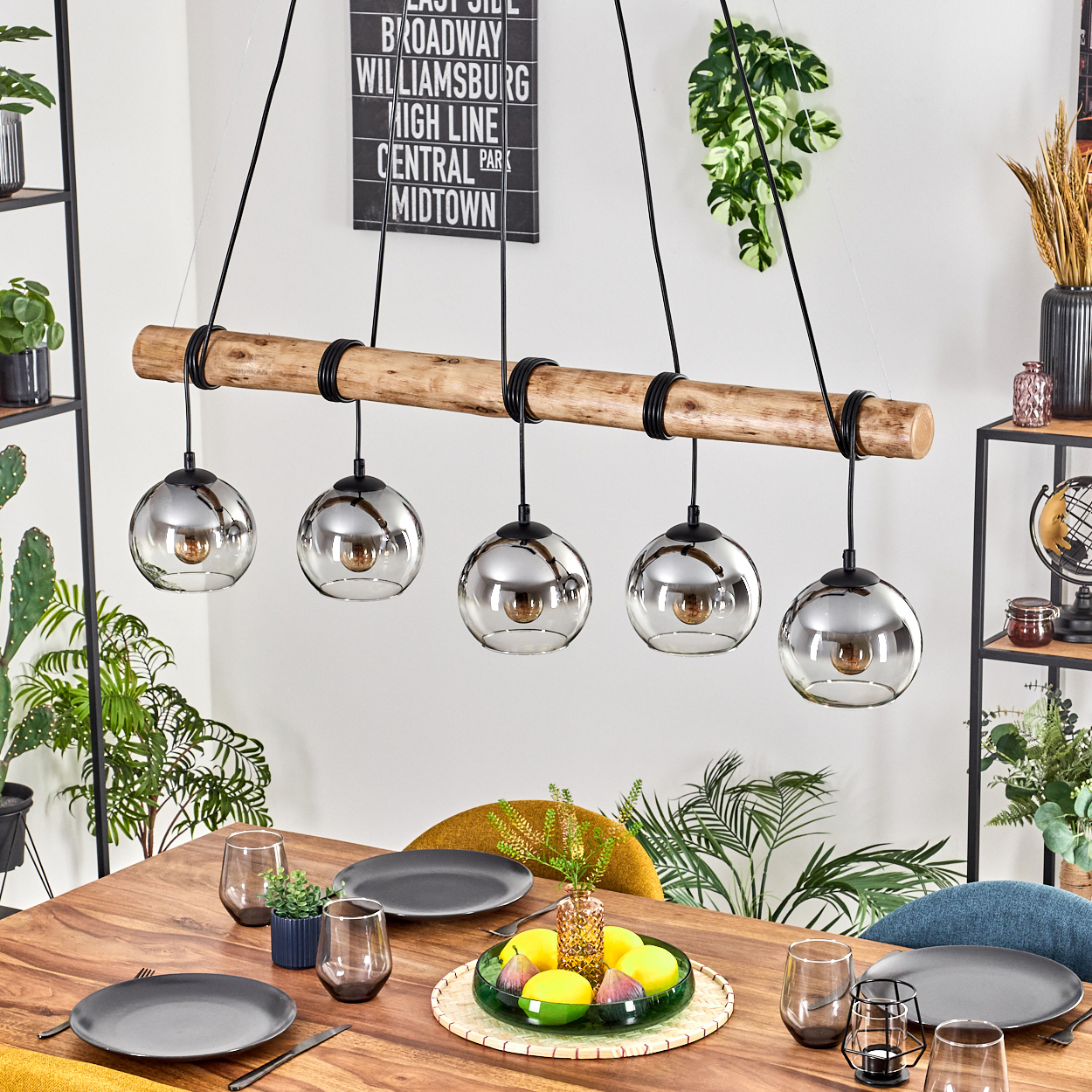 Esszimmer mit Holztisch, Tellern, Gläsern und Kronleuchter mit fünf Glaskugeln. Hintergrund: Pflanzen und ein Schild.