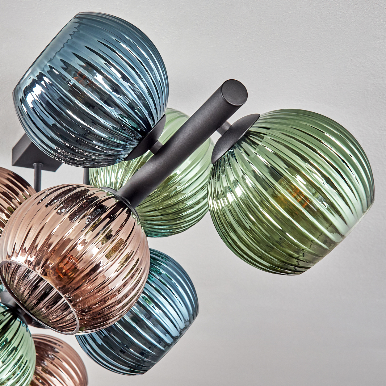 Nahaufnahme einer modernen Deckenleuchte mit farbigen Glaskugeln. Kugeln sind blau, grün und kupferfarben. Schwarze Metallarme halten sie vor weißem Hintergrund.