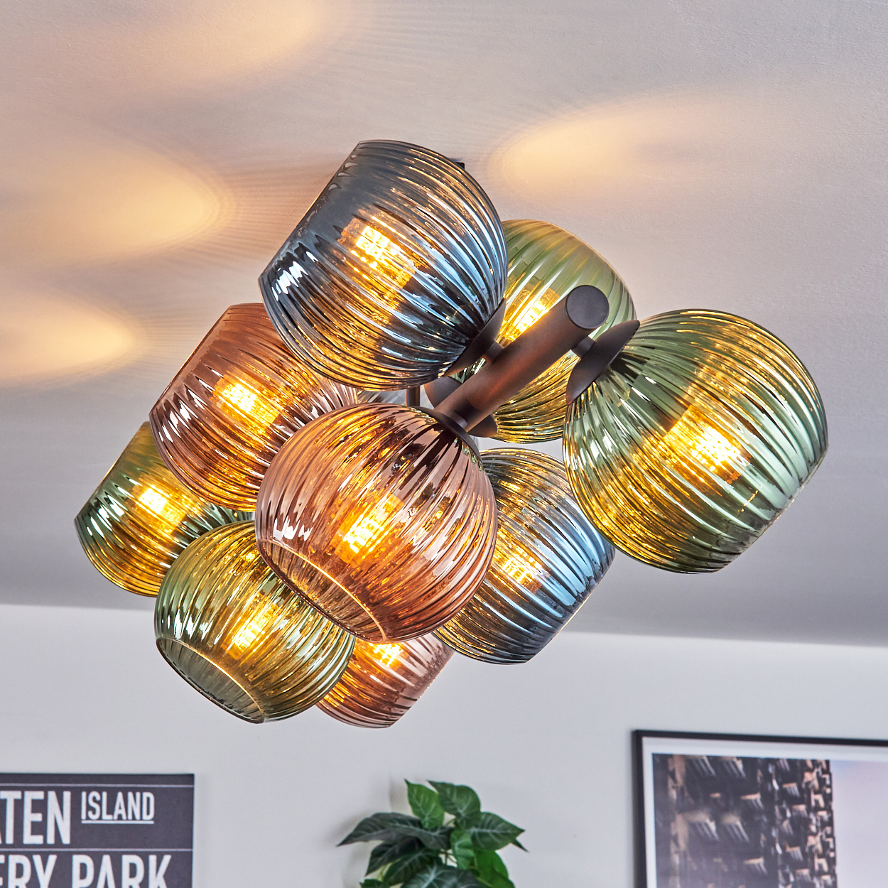 Eine moderne Deckenleuchte mit mehreren Glaskugeln in verschiedenen Farben. Die Lichter sind an und werfen ein warmes Licht. Eine Pflanze und gerahmte Kunst sind sichtbar.