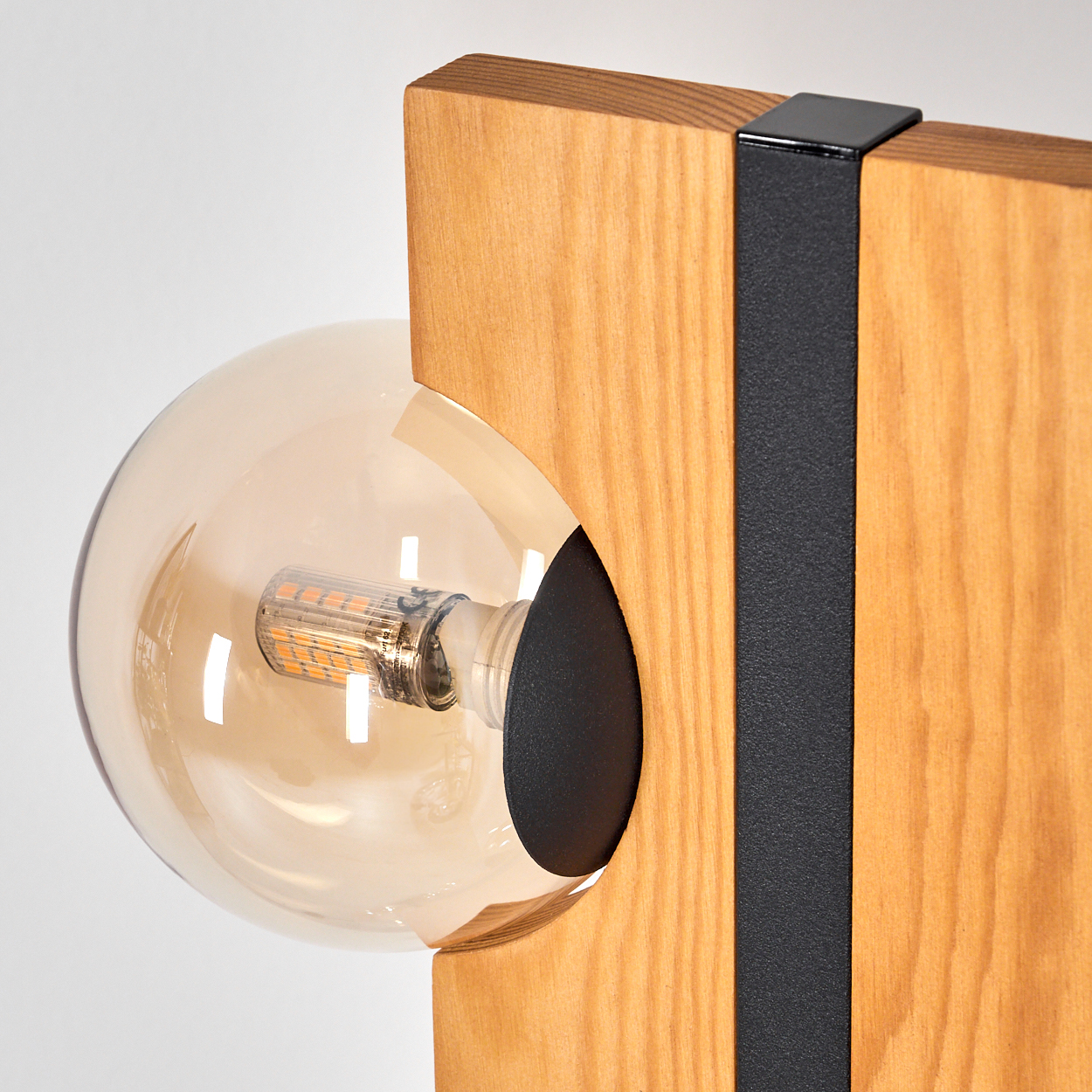 Nahaufnahme einer hölzernen Lampe mit einer runden, bernsteinfarbenen Glühbirne. Schwarze Metallakzente rahmen das Holz ein. Weißer Hintergrund.