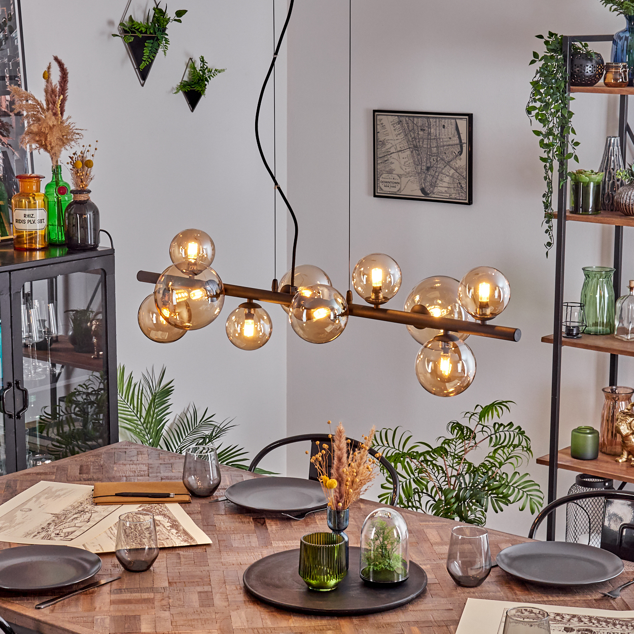 Esszimmer mit Holztisch, modernem Kronleuchter, Tellern, Gläsern und Pflanzen. Eine Karte hängt an der Wand.