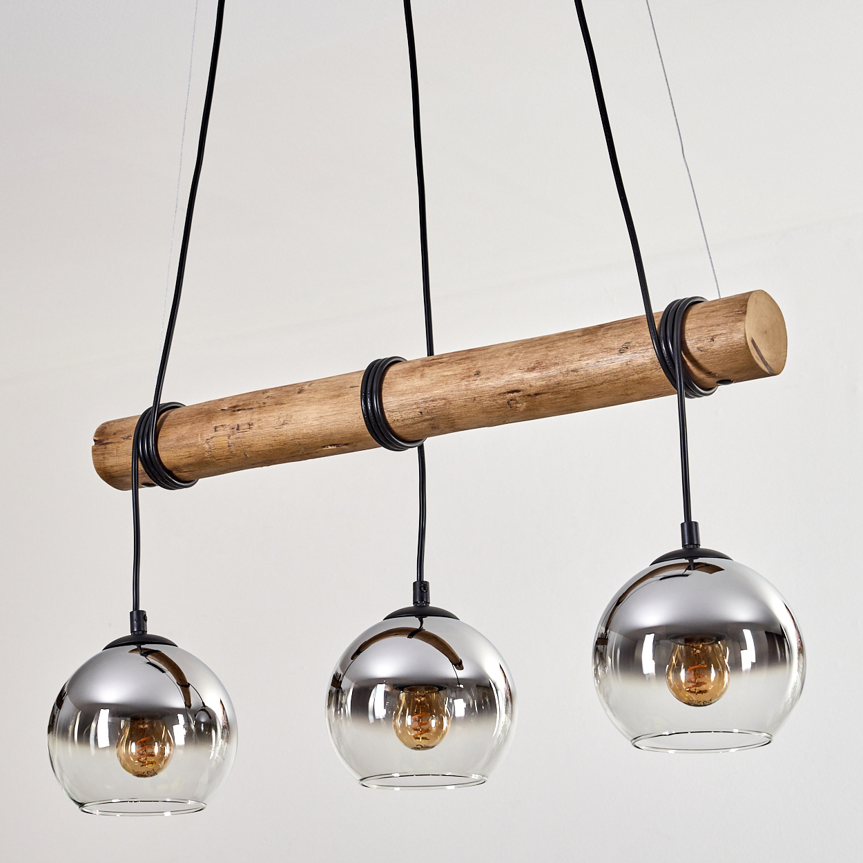 Eine moderne Pendelleuchte mit drei Glaskugeln, die an einem Holzbalken hängen. Jede Kugel beherbergt eine Glühbirne. Der Hintergrund ist weiß.
