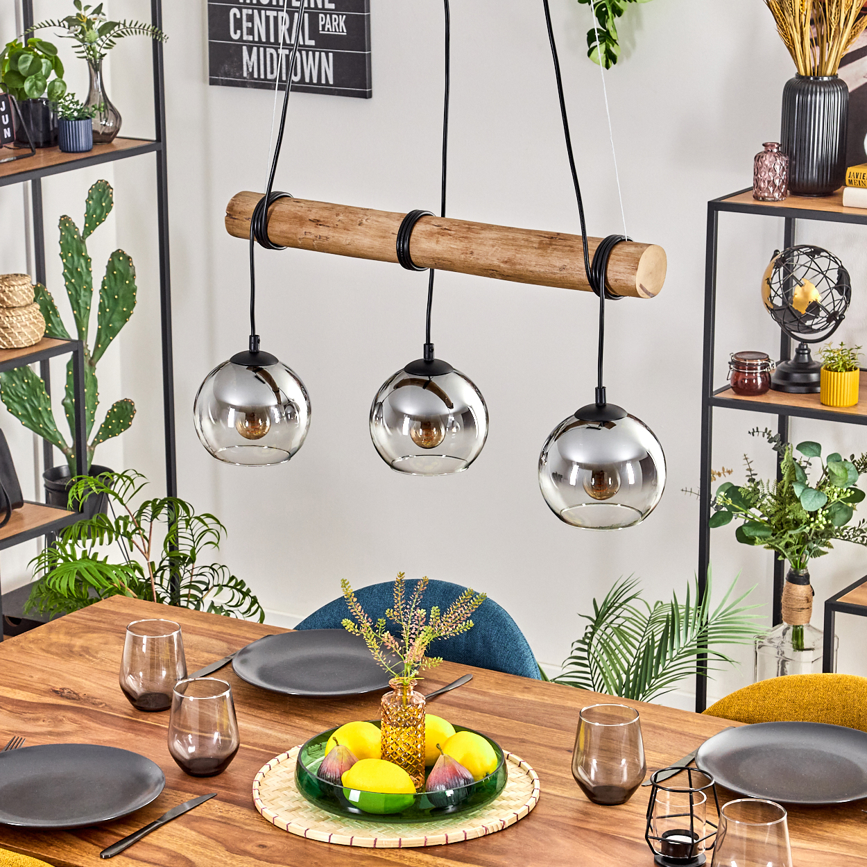 Ein Esstisch mit Tellern, Gläsern und einem Obst-Mittelstück. Oben hält ein Holzbalken drei Pendelleuchten.