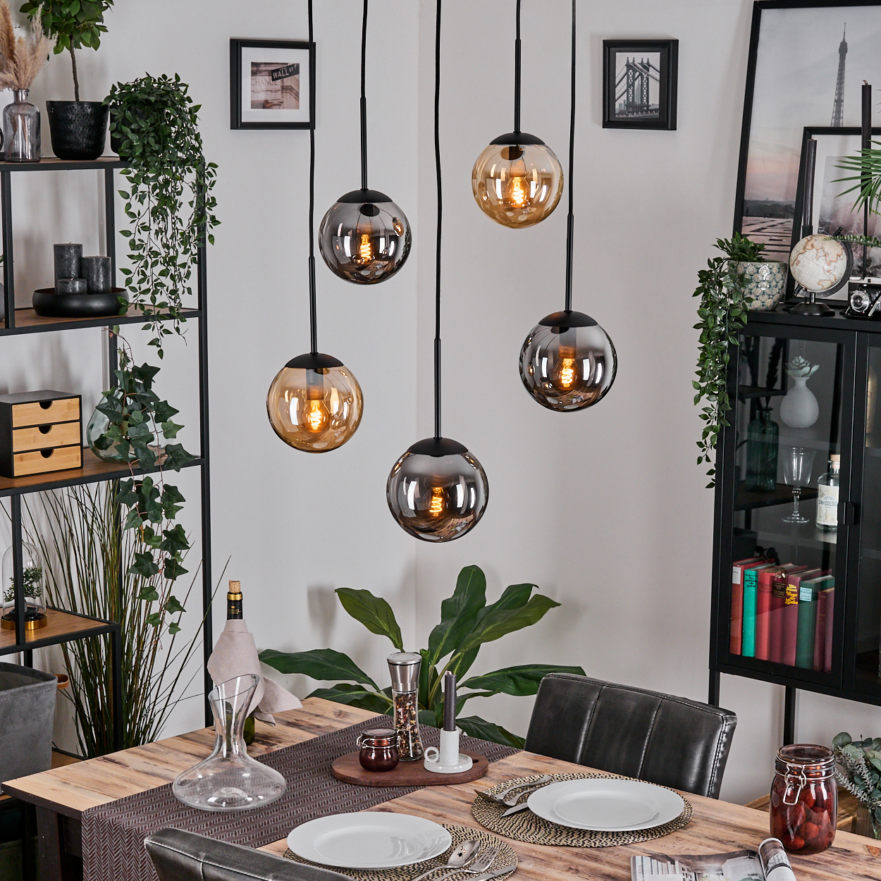 Esszimmer mit Holztisch, Glaskugelleuchten, Pflanzen und schwarz gerahmten Bildern. Tisch mit Tellern und Besteck gedeckt.