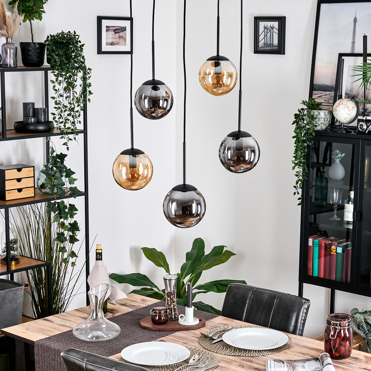 Esszimmer mit Holztisch, Hängelampen und Pflanzen. Teller und Besteck. Schwarze Regale mit Dekor.