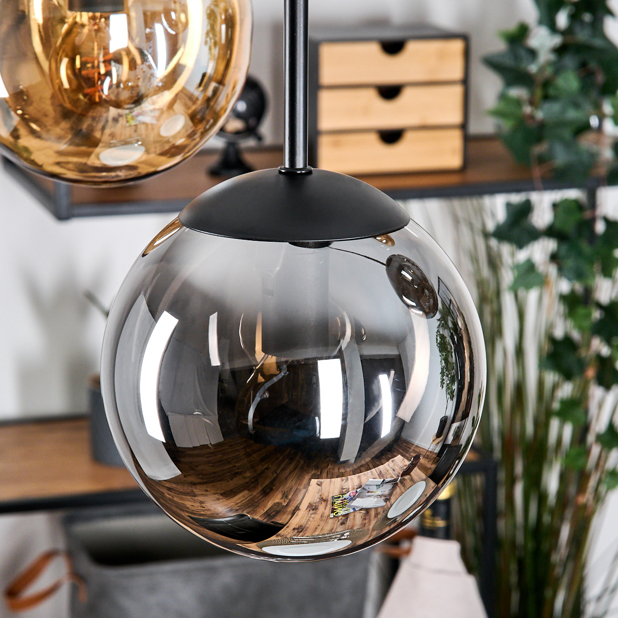 Nahaufnahme von zwei Glaskugel-Pendelleuchten, eine grau und eine goldfarben, die den Raum reflektieren. Regale und Pflanzen im Hintergrund.