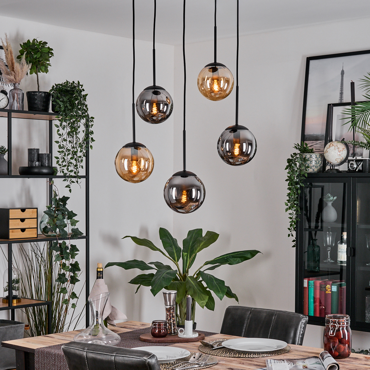 Esszimmer mit Holztisch, hängenden Kugelleuchten, Pflanzen und einem Schrank mit Dekorationen. Teller und Gläser stehen auf dem Tisch.