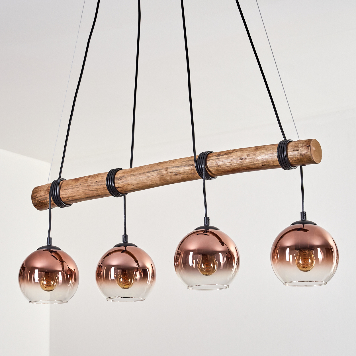 Eine moderne Leuchte mit vier kupferfarbenen Glaskugelleuchten, die an einem Holzbalken hängen. Es sind auch schwarze Kabel und Ringe vorhanden.
