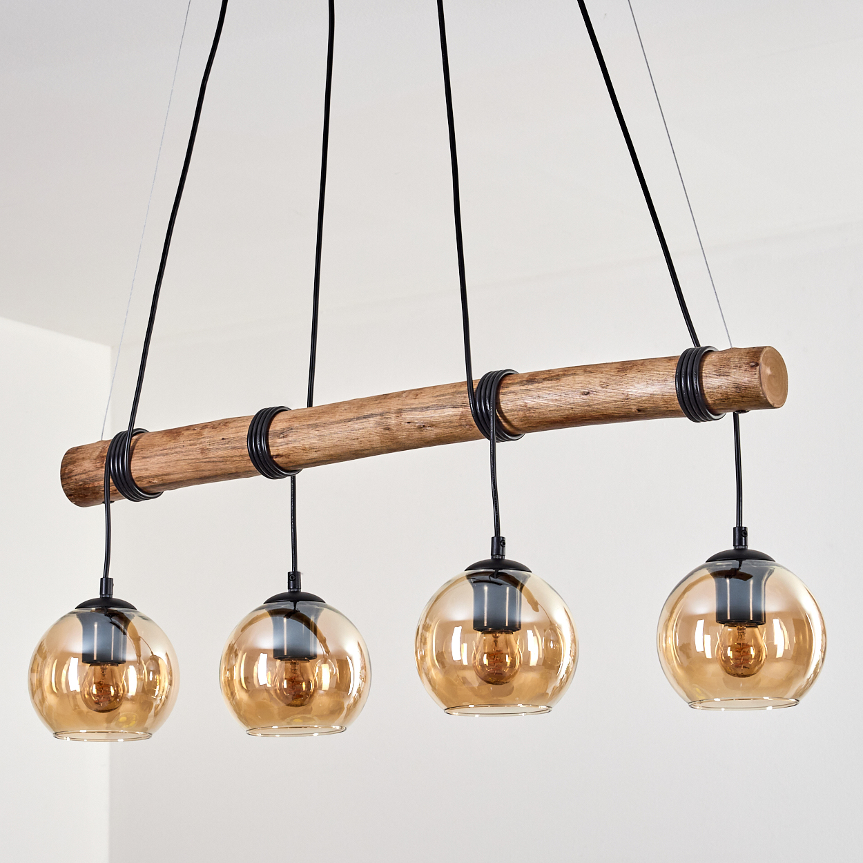 Ein Holzbalken mit vier Hängeleuchten. Jede Leuchte hat eine braune Glaskugel und schwarze Halterungen, vor einer weißen Wand.