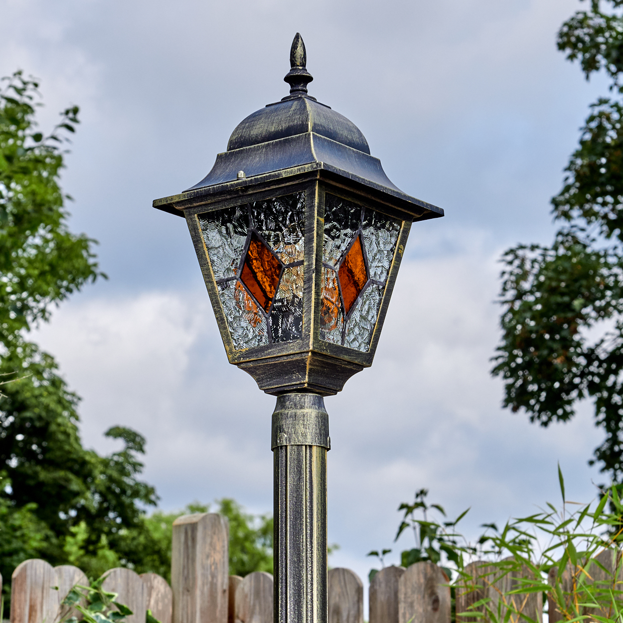 Eine dekorative Außenlampe mit Buntglasscheiben. Sie hat eine bronzefarbene Oberfläche, vor einem bewölkten Himmel und einem Holzzaun.