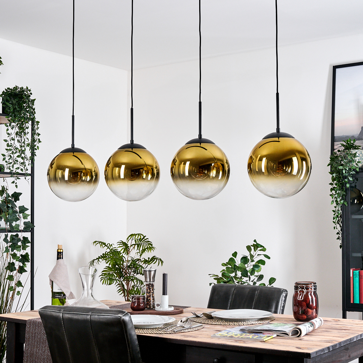 Vier goldene Pendelleuchten hängen über einem Esstisch mit Pflanzen und Dekorationen. Schwarze Stühle und ein schwarzer Schrank sind sichtbar.