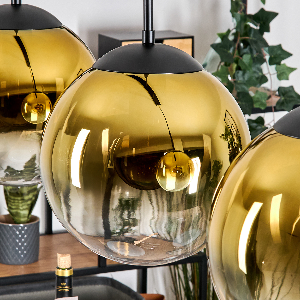 Drei goldene, kugelförmige Pendelleuchten hängen von der Decke. Im Hintergrund steht eine Pflanze in einem grauen Topf. Reflexion sichtbar.