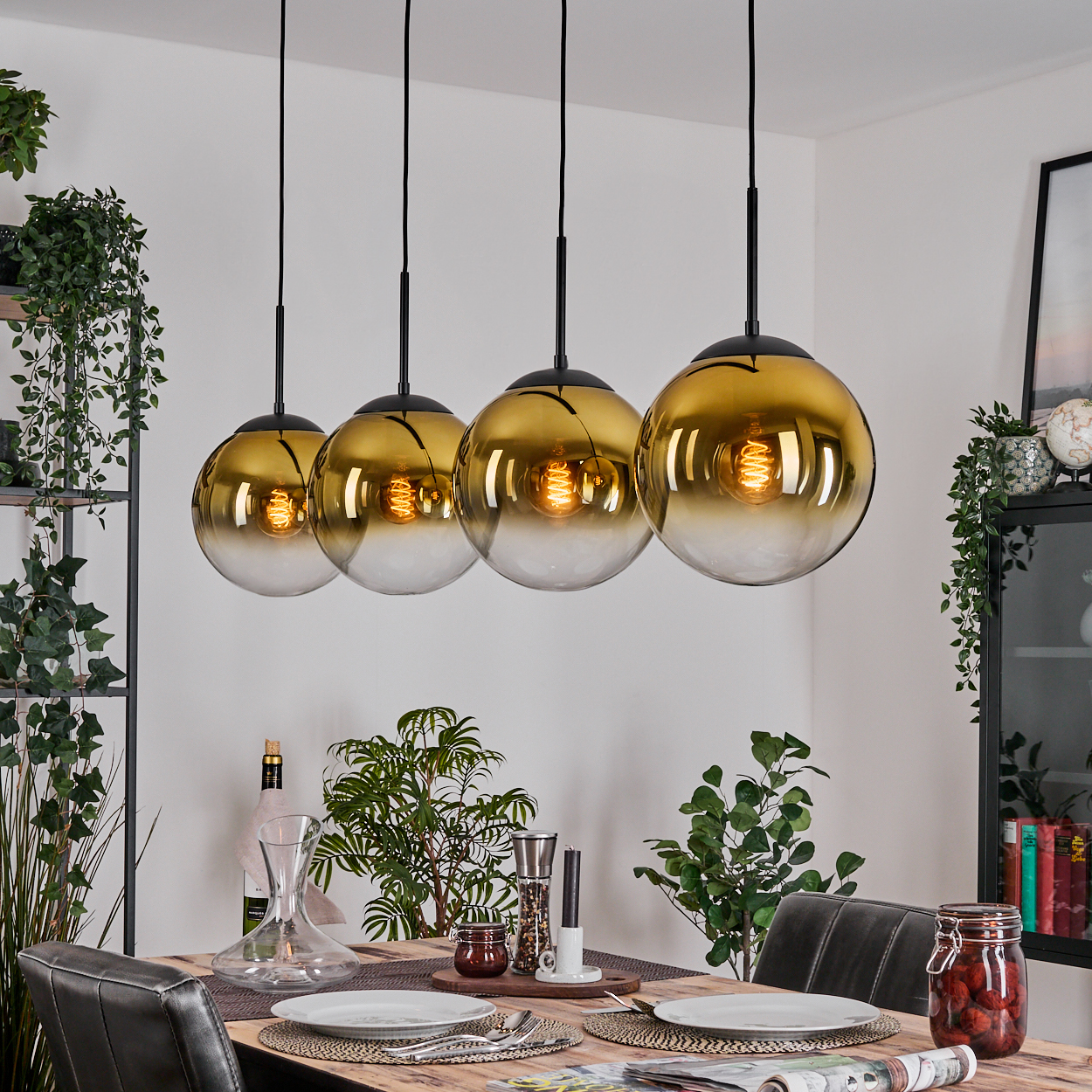 Vier goldfarbene Pendelleuchten über einem Esstisch, der für das Abendessen gedeckt ist. Pflanzen und dekorative Gegenstände umgeben den Tisch.