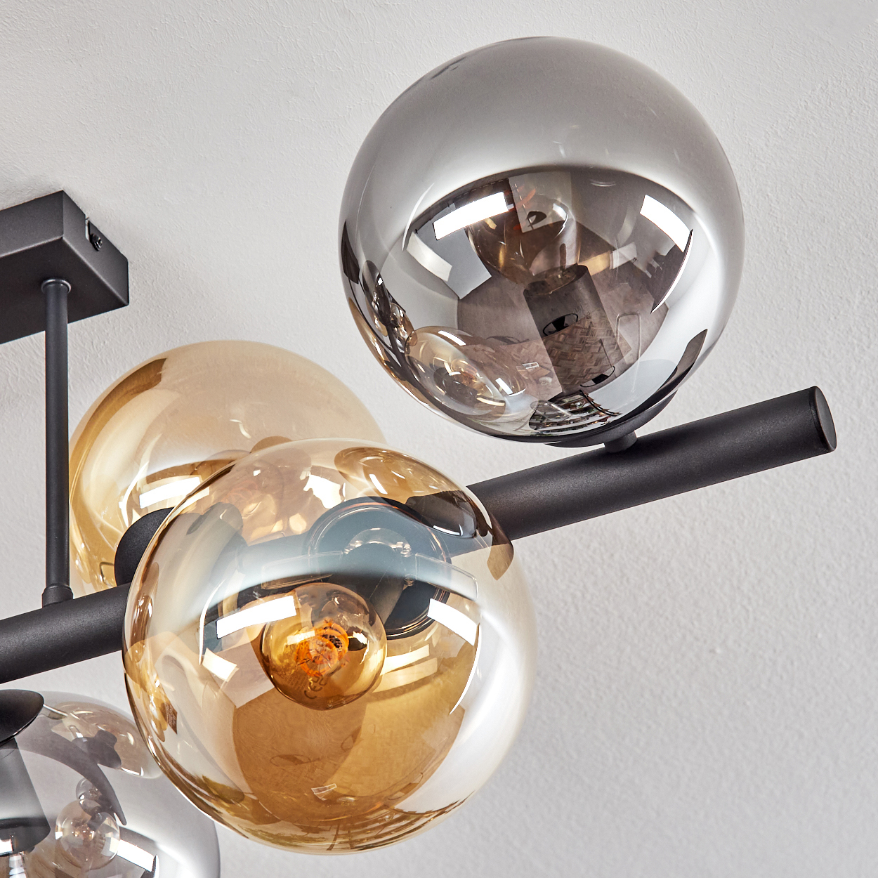 Moderne Deckenleuchte mit kugelförmigen Glasschirmen. Einige sind klar, andere bernsteinfarben oder silber, montiert auf schwarzen Stangen, vor einer weißen Wand.