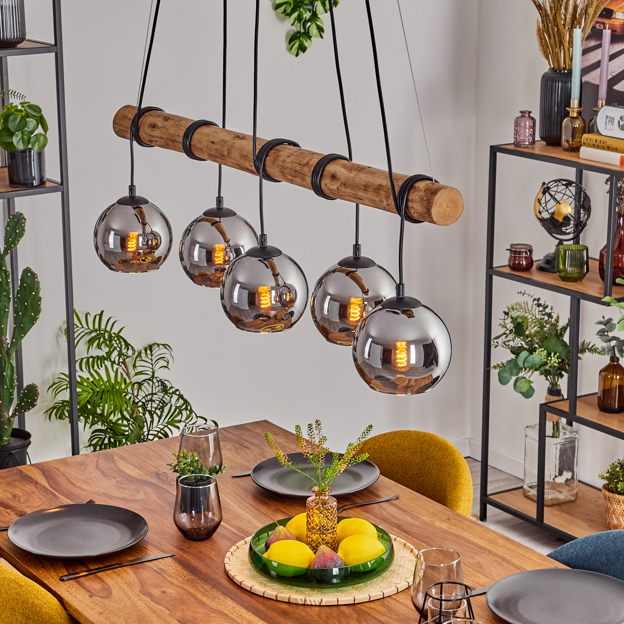 Esszimmer mit Holztisch, Kronleuchter und Tischarrangement mit Obst. Regale mit Pflanzen und Dekoration im Hintergrund.