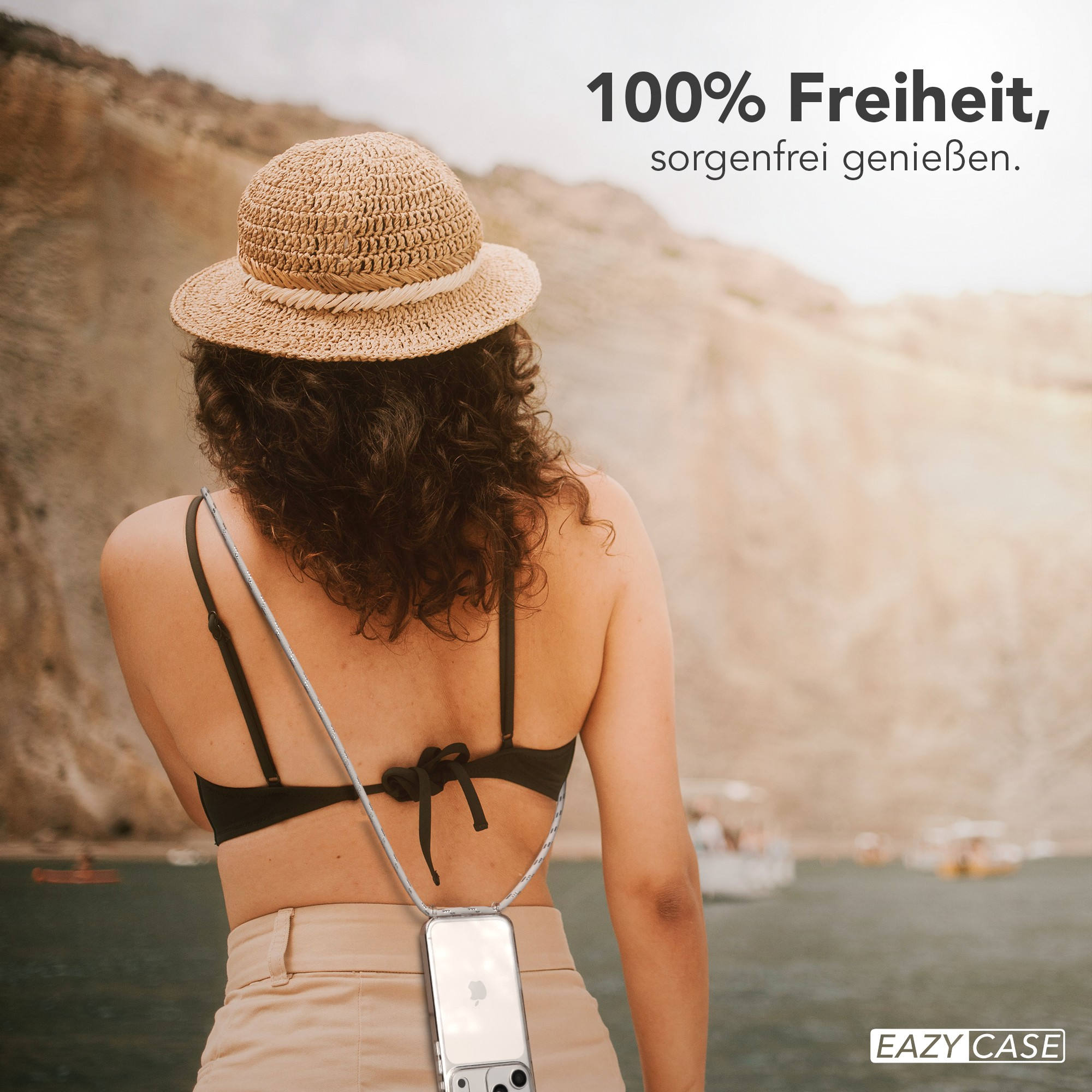 Frau mit Strohhut, schwarzem Oberteil und beiger Hose. Eine Handyhülle hängt um ihren Hals, mit Wasser und Bergen im Hintergrund.