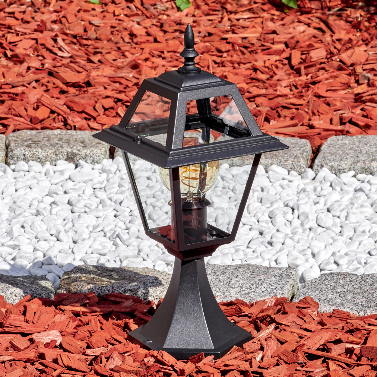 Schwarze Außenlampe mit Glasscheiben, auf roter Mulch, weißen Steinen und grauem Steinrand. Warme Glühbirne.