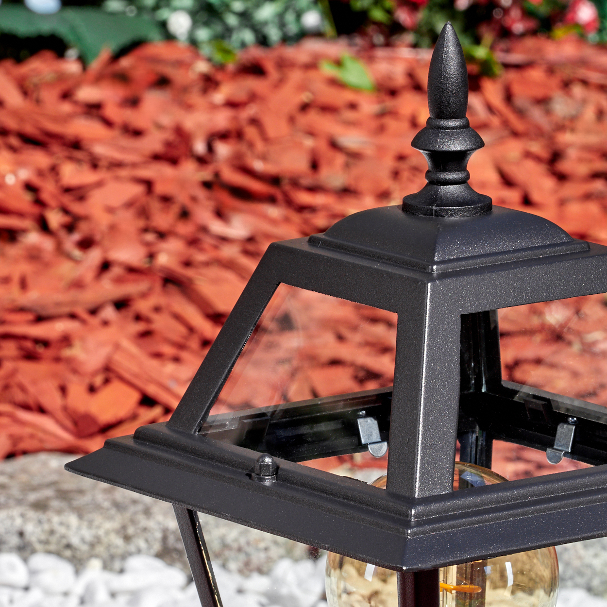 Eine schwarze Außenlampe mit Glasgehäuse steht auf einer weißen Steinfläche, im Hintergrund roter Mulch.