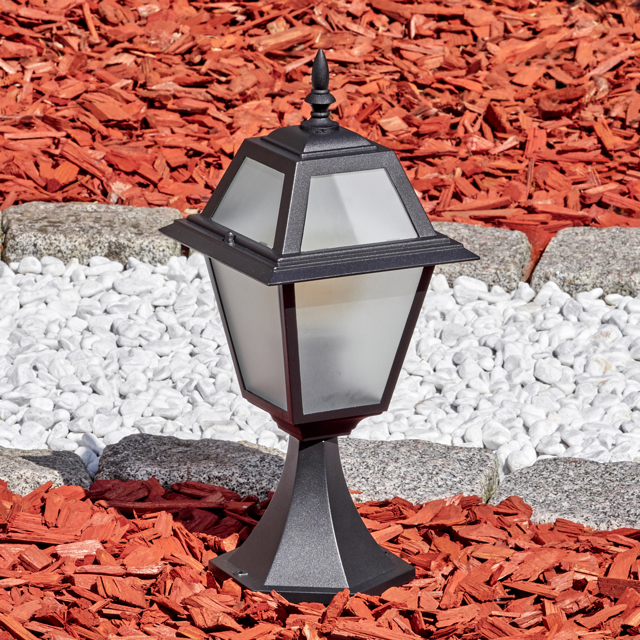 Schwarze Außenlampe auf roter Mulch. Weiße Steine und graue Steine umranden den Sockel der Lampe.