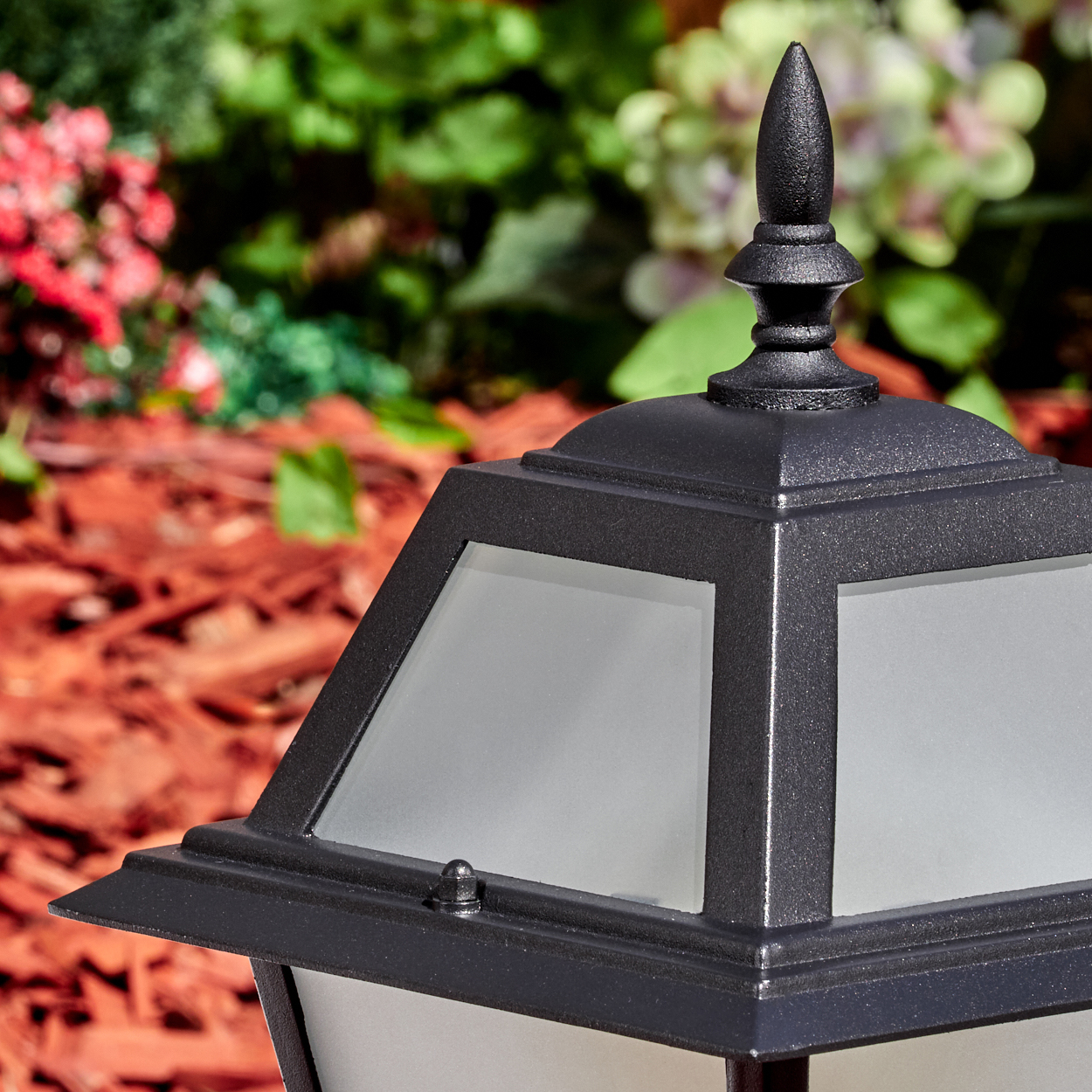 Nahaufnahme einer schwarzen Außenlampe mit Milchglasscheiben. Im Hintergrund roter Mulch, grüne Pflanzen und Blumen.