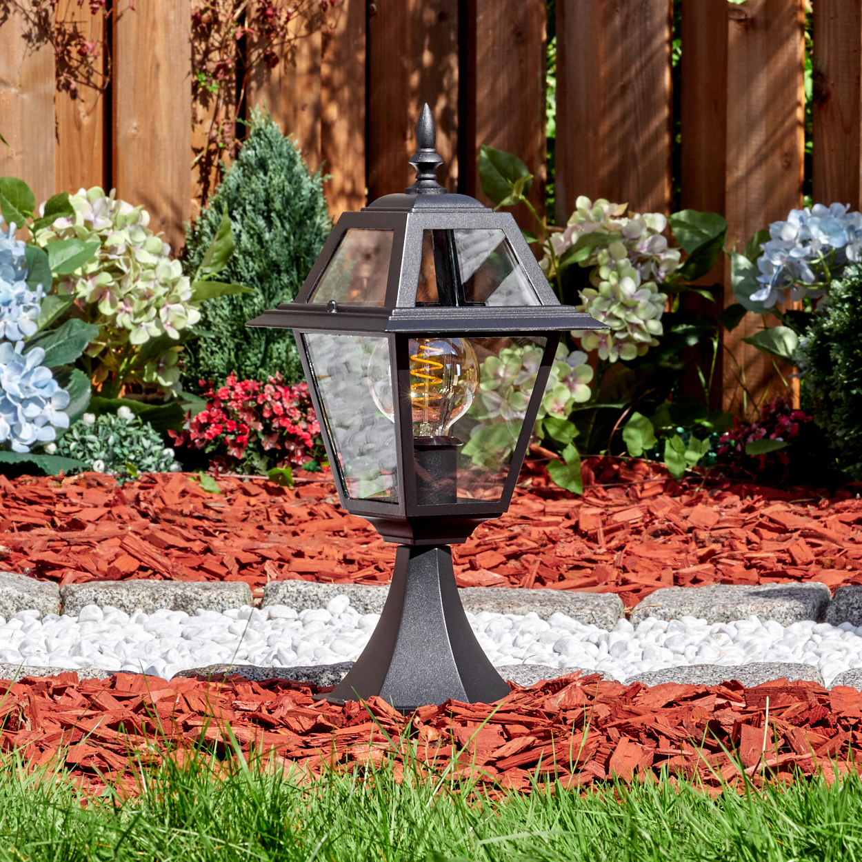 Eine schwarze Außenlampe mit Glasgehäuse steht in einem Garten. Der Garten hat Holzschnitzel, Blumen und einen Holzzaun.