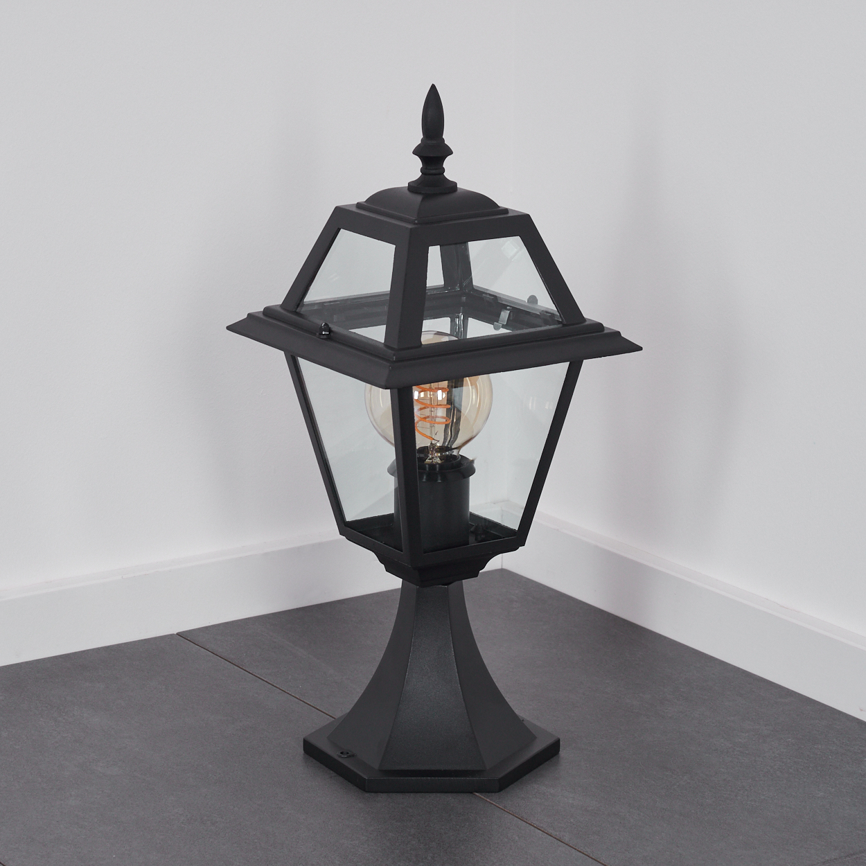 Eine schwarze Außenlampe mit Glasgehäuse und dekorativer Spitze, steht auf einem dunkelgrauen Boden und vor einer weißen Wand.