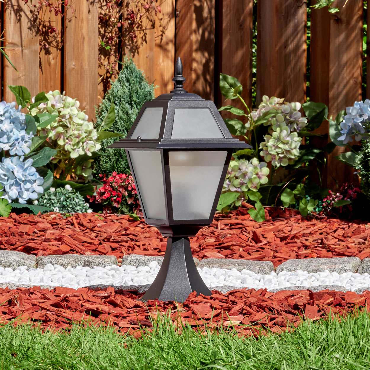 Eine schwarze Außenlampe steht in einem Gartenbeet mit roter Mulche und weißen Steinen. Blumen und ein Holzzaun sind im Hintergrund.